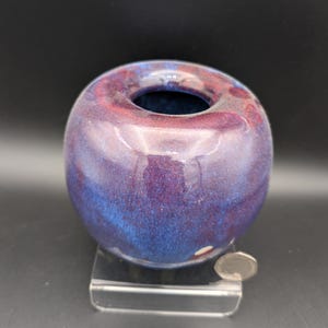 Könnte beinhalten: Kleine, runde Keramikvase mit glänzendem Finish. Die Vase hat eine Mischung aus Farben, darunter tiefes Blau, Lila und rotbraune Farbtöne. Die Öffnung ist dunkelblau. Die Vase wird auf einem transparenten Acrylständer präsentiert.