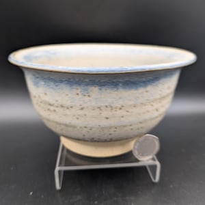 May include: A ceramic bowl with a wide, rounded shape. The bowl has a light beige base with a blue rim and speckled detailing. It sits on a clear plastic stand, with a coin for scale. The bowl is likely handmade and suitable for serving or display.