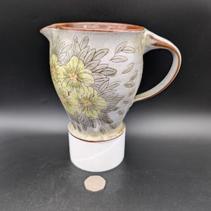 May include: A ceramic pitcher with a floral design in shades of green and yellow. The pitcher has a brown rim and a white base.
