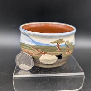 May include: A small, hand-painted ceramic bowl with a landscape design. The bowl features a brown interior, a cream-colored exterior with a painted scene of trees, a sheep, and a blue sky. The bowl is approximately 3 inches in diameter.