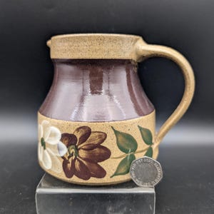 May include: A ceramic pitcher with a brown and tan color scheme. The pitcher features a dark brown center section, a tan rim and handle, and a floral design with white and brown flowers and green leaves. The pitcher has a curved handle.