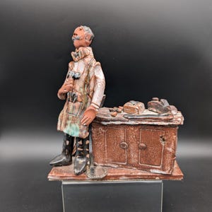 Peut inclure: Figurine en céramique représentant un homme en manteau long appuyé contre un meuble marron. La figurine a une moustache et porte des bottes noires. Le meuble a deux portes et divers objets sur le dessus. La palette de couleurs comprend le marron, le blanc et des touches de vert.