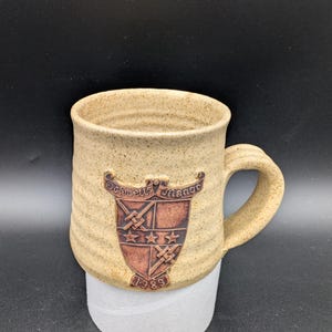 May include: A tan ceramic mug with a brown crest featuring the text "Schwells Manor" and the year "1989".