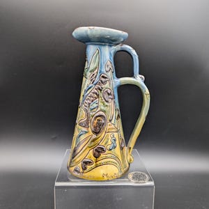 May include: A vintage ceramic pitcher with a blue handle and spout, and a yellow base. The pitcher is decorated with a floral design in shades of blue, green, and brown. The pitcher is displayed on a clear acrylic stand.