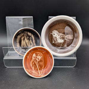 Op de afbeelding: Drie kleine, ronde keramische schalen met bruine en witte ontwerpen. Eén schaal toont een vogel, een andere een uil en de derde figuren. De uilenschaal heeft de tekst "Norfolk Wildlife Park". De schalen worden tentoongesteld op een transparante plastic standaard.