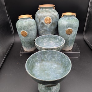 Può includere: Una collezione di vasi e ciotole in ceramica fatti a mano nei toni del blu e del verde. I vasi hanno una superficie strutturata, bordi dorati e medaglioni decorativi. Le ciotole sono rotonde, di cui una su un piedistallo.