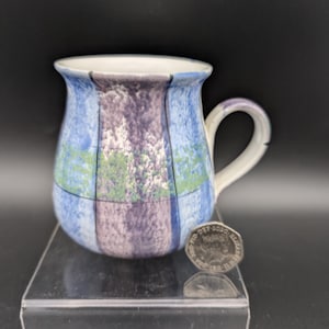 May include: A ceramic mug with a rounded body and a handle. The mug features a design of vertical panels in blue, purple, and green, with a white interior and handle. A coin is next to the mug.