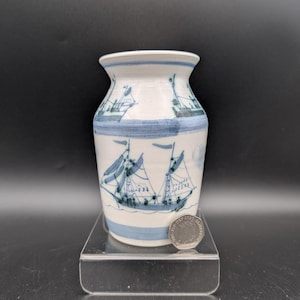 May include: A white ceramic vase decorated with blue sailboat motifs. The vase features a flared rim and blue bands at the top and base. The sailboat design is repeated around the body, creating a nautical theme. The vase is approximately 15 cm tall.