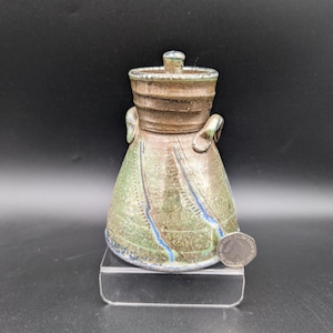 Może przedstawiać: Ręcznie robiony ceramiczny słoik z pokrywką. Słoik jest mieszanką odcieni zieleni, brązu i błękitu, z teksturowaną powierzchnią. Ma dwa małe uchwyty i mały guziczek na pokrywce. Słoik ma około 10 cm wysokości.