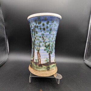 Könnte beinhalten: Eine handgefertigte Keramikvase mit ausgestellter Form. Die Vase hat ein blau-grünes Sprenkelmuster mit einer Landschaft von Bäumen. Der Rand ist weiß und der Boden hellbraun. Die Vase ist etwa 15 cm hoch.