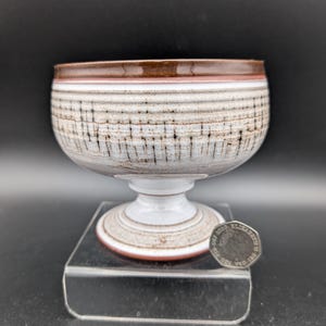 Peut inclure: Un bol en céramique sur pied avec un bord marron et un motif rayé blanc et gris. Le bol a un socle et est exposé sur un présentoir en acrylique transparent. Une pièce de monnaie est à côté du bol.