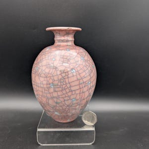 May include: A pink ceramic vase with a dark blue crackle glaze pattern and a small flared rim. The vase is positioned next to a silver-coloured coin for scale, which measures approximately 2.5 cm in diameter, providing a clear visual reference for size.