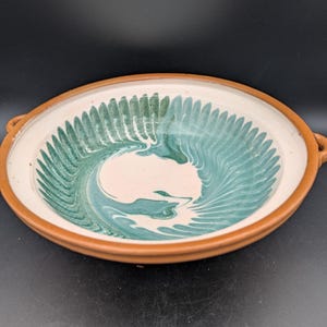 May include: A handmade ceramic bowl with a unique design. The bowl features a white interior with a teal and green swirled pattern. The rim and handles are a warm brown color. The bowl is perfect for serving food.