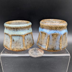 Peut inclure: Deux tasses en céramique faites à la main avec un design hexagonal unique. Les tasses présentent un mélange de teintes bleues, marron et beige, avec une bande bleu clair au milieu. Elles sont présentées sur une surface transparente.