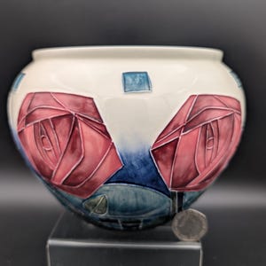 Könnte beinhalten: Eine weiße Keramikschale mit breiter Öffnung, verziert mit zwei großen roten Rosen mit blauen und grünen Akzenten. Die Schale wird auf einer schwarzen Oberfläche präsentiert.
