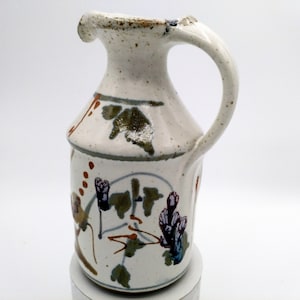 Könnte beinhalten: Ein kleiner weißer Keramik Krug mit braunem Henkel und einem Blumendesign in Braun-, Grün-, Blau- und Violetttönen.