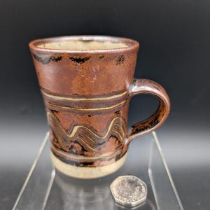 Peut inclure: Une tasse en céramique faite à la main avec un glaçage marron et un motif ondulé marron foncé et beige. La tasse a une anse et une base beige clair. Une pièce de monnaie est à côté de la tasse.