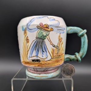 Può includere: Una tazza in ceramica dipinta a mano con l'illustrazione di una persona con un cappello che raccoglie il grano. La tazza ha un manico e una base turchese, con un corpo bianco e crema. L'opera d'arte presenta toni blu, verdi e marroni.