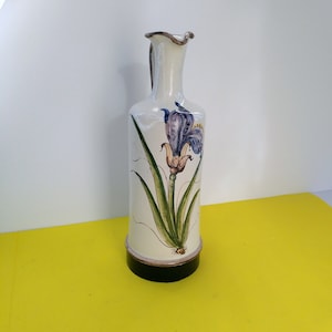May include: A white ceramic vase with a brown rim and a black base. The vase is decorated with a hand-painted floral design of a purple iris with green leaves.