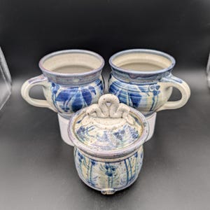 May include: Three handmade ceramic mugs with blue and white floral designs. The mugs have a rounded shape and a handle. One mug has a lid with a decorative knob.