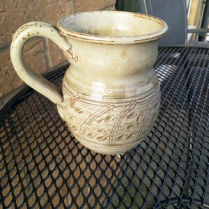 May include: A light green ceramic mug with a handle and a decorative design of leaves. The mug is sitting on a black metal table.