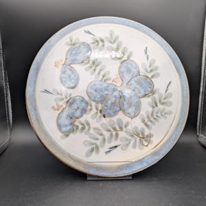 May include: Circular ceramic plate with a white centre and a blue rim. The plate features a floral design with blue and green leaves. The plate is a decorative piece for serving or display.