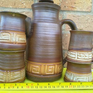 May include: A set of four brown pottery mugs with a geometric design. The mugs are stacked on top of each other, with the largest mug at the bottom. The mugs are all decorated with a brown and white geometric design. The mugs are sitting on a yellow surface.