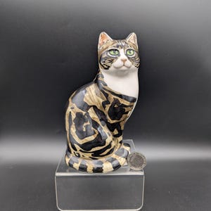 Peut inclure: Figurine de chat en céramique, en position assise. Le chat a un visage et une poitrine blancs, des yeux verts et un pelage à motifs noirs et beiges. Présenté sur un présentoir en acrylique transparent sur fond noir.