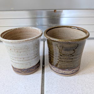 May include: Two handmade ceramic cups. One is off-white with brown accents, the other is dark brown. Both have a decorative band near the base and a textured surface. The cups are likely for drinking.