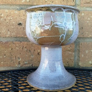 May include: A ceramic goblet with a brown and white glaze. The goblet has a wide base and a round bowl. It is sitting on a black metal table.
