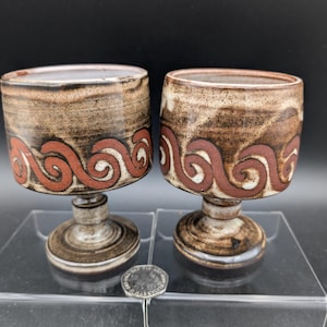 May include: Two ceramic goblets in brown and cream tones. Each goblet has a decorative red and white swirl pattern near the rim. The goblets are displayed on a clear surface against a dark backdrop.