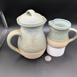 May include: A set of two ceramic pitchers with a green and brown glaze. The larger pitcher has a lid and a handle. The smaller pitcher has a handle.