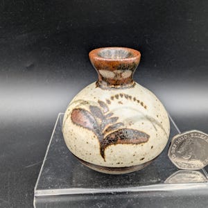 May include: A small, round ceramic vase with a speckled off-white glaze and a dark brown, fern-like botanical pattern. The vase features a narrow neck with a thick, dark brown rim. A British twenty pence coin is placed next to the vase for scale.