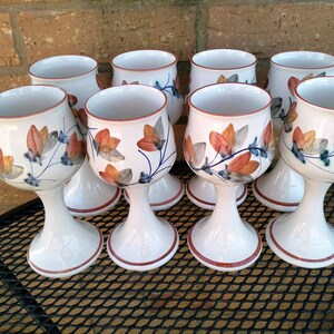 May include: Set of eight white ceramic goblets with a floral design in shades of orange, brown, blue, and green. The goblets have a wide bowl and a tall, slender stem.