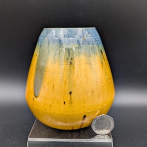 Puede incluir: Un jarrón de cerámica hecho a mano con una base ancha y una parte superior estrecha. El jarrón presenta un esmalte degradado de colores, que va del azul en la parte superior al amarillo en el medio y un amarillo más oscuro en la parte inferior. Pequeñas motas negras salpican la superficie.