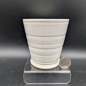 May include: A white ceramic vessel with a tapered cylindrical shape, featuring a beaded rim and base, and horizontal bands of diagonal herringbone texture. A British twenty pence coin is placed next to the base for scale reference.