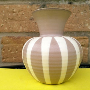 Peut inclure: Un vase en céramique à rayures, avec un corps arrondi et un col évasé. Les rayures verticales alternent le beige et le blanc. Le fond est un mur de briques et une surface jaune.