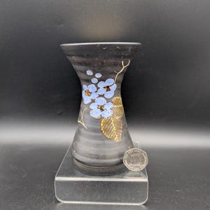 May include: A black ceramic vase with a flared rim and a tapered body, featuring a hand-painted design of light blue flowers and green leaves. A silver-colored coin is placed next to the base for scale, showing the size of the object.