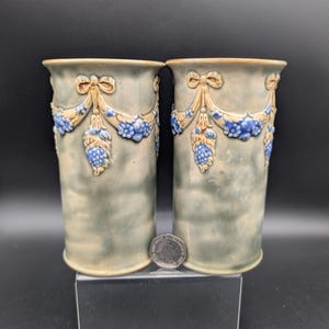 May include: Two cylindrical ceramic vases with a light green and blue glaze. Each vase features a decorative relief of blue flowers and tan bows. The vases are displayed on a clear stand against a black background.