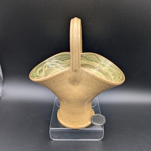 May include: A ceramic basket with a handle, featuring a tan exterior and a green and tan patterned interior. The basket is displayed on a clear stand against a dark background. The basket's design is reminiscent of a flower.
