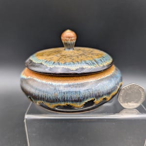 May include: A small, handcrafted ceramic jar with a lid. The jar is primarily blue, brown, and orange with a glossy finish. The lid has a small knob on top. The jar is sitting on a clear surface.