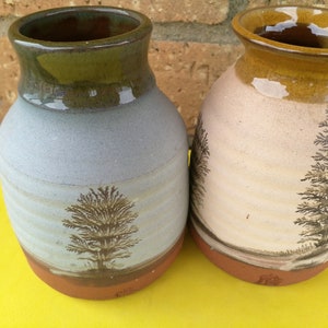 May include: Two ceramic vases with brown, white and blue glaze. Each vase has a tree design painted on the front. The vases are sitting on a yellow surface.