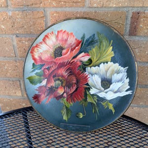 May include: Circular decorative plate with a floral design. It features red, white, and dark red poppies against a dark teal background, with a gold-coloured rim. Green leaves and a small bud are also visible.