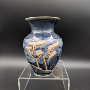 Peut inclure: Un vase en céramique fait à la main avec un glaçage bleu foncé et des motifs en forme d'arbres beiges. Le vase a un corps arrondi et un bord évasé. Le vase est exposé sur un présentoir en acrylique transparent. Une pièce de monnaie est à côté du vase.