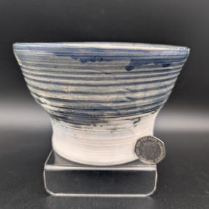 Pode incluir: Uma tigela de cerâmica artesanal com um design listrado azul e branco. A tigela tem uma parte superior larga e uma base mais estreita. O exterior apresenta listras horizontais em tons de azul e branco. A tigela é exibida em um suporte transparente.