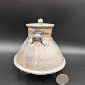 May include: A handmade ceramic jar with a lid. The jar is a light gray color with a textured surface and a flared base. It has a blue rim and handle accents. The lid has a small blue knob. The jar is approximately 4 inches tall.