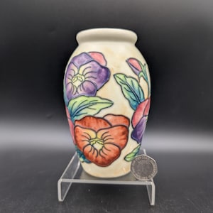 Op de afbeelding: Een kleine, crèmekleurige keramische vaas met een bloemenontwerp. De vaas heeft handgeschilderde bloemen in tinten paars, rood en oranje, met groene bladeren. De vaas wordt tentoongesteld op een transparante acrylstandaard.