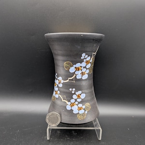 May include: A matte black hourglass-shaped vase featuring a hand-painted floral pattern with light blue petals, brown centers, and gold-outlined leaves. A silver-colored coin is placed at the base for scale, resting on a clear acrylic stand.