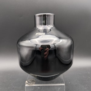 May include: A black glass vase with a rounded body and a narrow neck. The vase is displayed on a clear acrylic stand. The glossy surface reflects light, creating highlights and shadows. The vase has a modern, minimalist design.