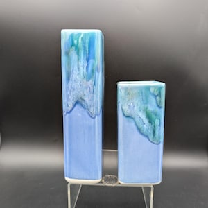 May include: Two rectangular ceramic vases. The vases are a light blue color with a darker blue and green glaze at the top, resembling a wave. The vases are displayed on a clear acrylic stand. The taller vase is approximately 10 inches tall, and the shorter vase is approximately 6 inches tall.